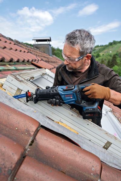 A person wearing safety equipment uses a reciprocating saw to cut wood on a rooftop.
