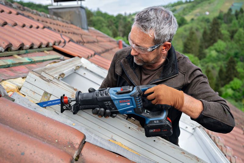 Person wearing safety equipment cuts wooden window frame on a roof with a reciprocating saw.