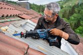 Person wearing safety equipment cuts wooden window frame on a roof with a reciprocating saw.