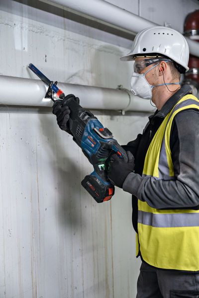 A person wearing safety equipment uses a reciprocating saw to cut a metal pipe.