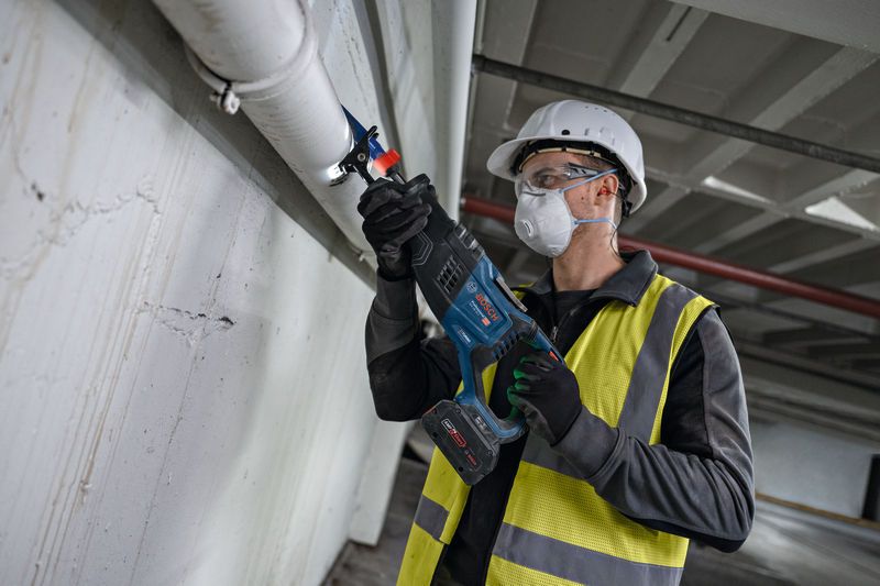 A person wearing safety equipment cuts a ceiling pipe using a reciprocating saw.