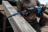 A person wearing safety equipment uses a reciprocating saw to cut a wooden beam.