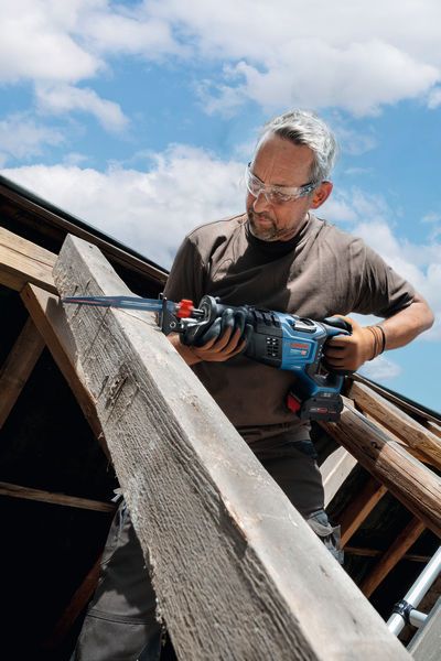 A person wearing safety equipment cuts a large wooden beam with a reciprocating saw.