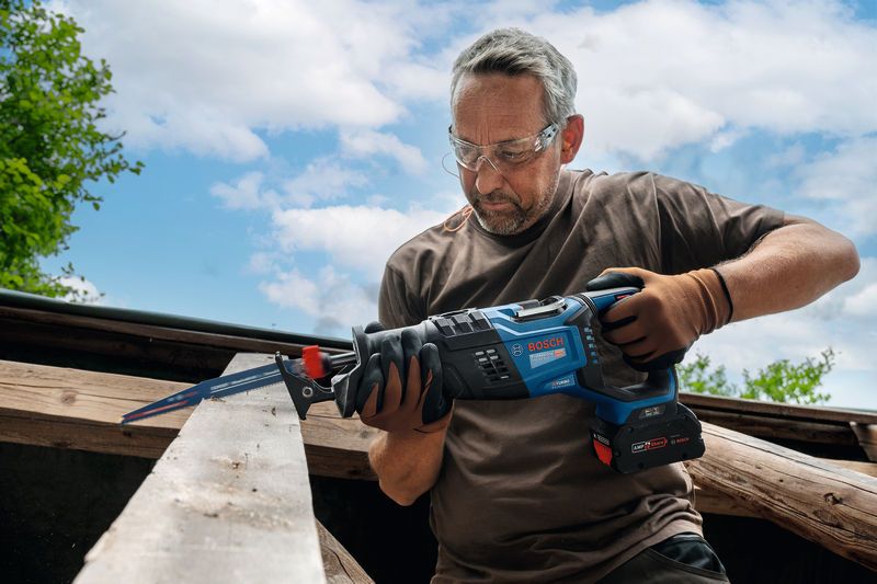 A person wearing safety equipment uses a reciprocating saw to cut a wooden beam outdoors.