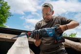 A person wearing safety equipment uses a reciprocating saw to cut a wooden beam outdoors.