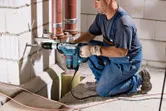A person wearing safety equipment drills into a concrete wall near pipes.