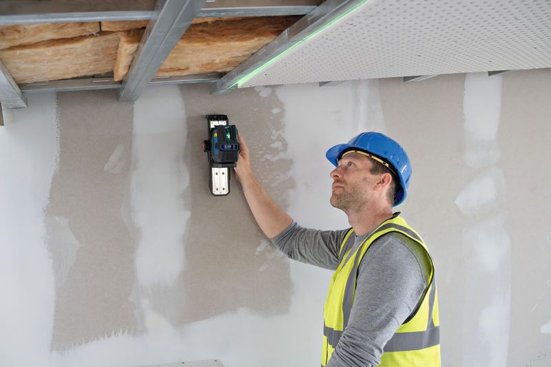 A person wearing safety equipment uses a laser leveling tool on a ceiling frame.