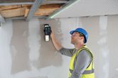 A person wearing safety equipment uses a laser leveling tool on a ceiling frame.