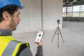 A person wearing safety equipment uses a smartphone to control a laser leveling tool on a tripod.