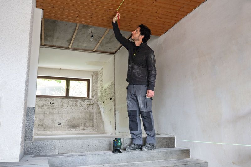 A person wearing safety equipment measures a ceiling using a laser leveling tool.