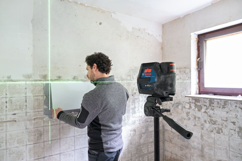 A person aligns tiles on a wall using a laser leveling tool on a tripod.