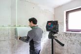 A person aligns tiles on a wall using a laser leveling tool on a tripod.