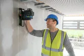 A person wearing safety equipment aligns a laser leveling tool on a wall.