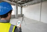 A person wearing safety equipment adjusts a laser leveling tool with a smartphone in a bare room.