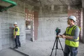 Two people wearing safety equipment set up a laser leveling tool on a tripod in a concrete room.