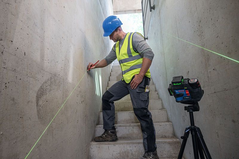 A person wearing safety equipment marks a wall using a laser leveling tool on stairs.