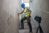 A person wearing safety equipment marks a wall using a laser leveling tool on stairs.
