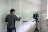 A person aligns tiles on a wall using a green laser leveling tool on a tripod.