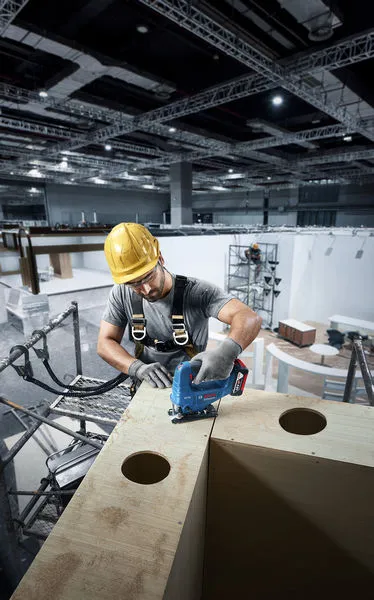A person wearing safety equipment uses a cordless jigsaw to cut wood on a construction site.