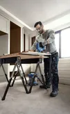 A person wearing safety equipment cuts wood with a cordless jigsaw on a workbench.