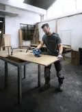 A person wearing safety equipment uses a cordless jigsaw to cut a wooden board.