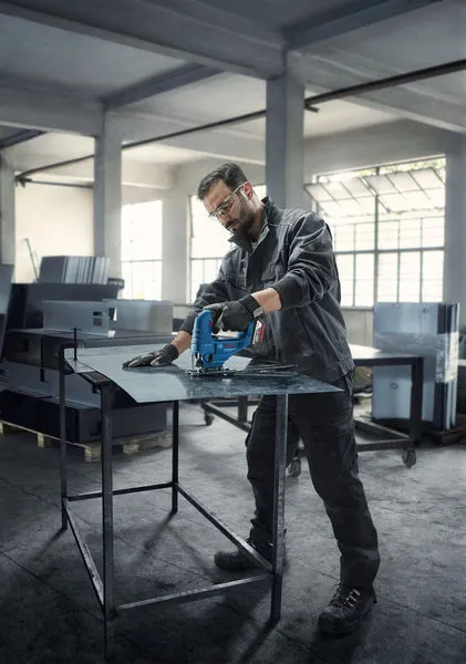 A person wearing safety equipment uses a cordless jigsaw to cut sheet metal in a workshop.