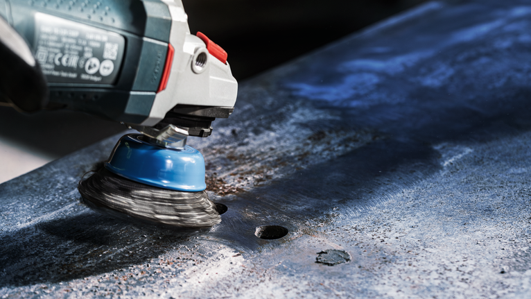 PRO Metal heavy Cup Brush, 75 x 0.5 mm, Thread A power tool with a wire cup brush grinds rust off a metal sheet.