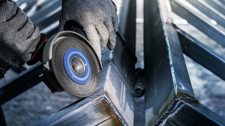 PRO Stainless Steel heavy X-Lock Wire Wheel, 115 x 0.5 mm Person wearing safety equipment grinds a metal beam with a power tool.