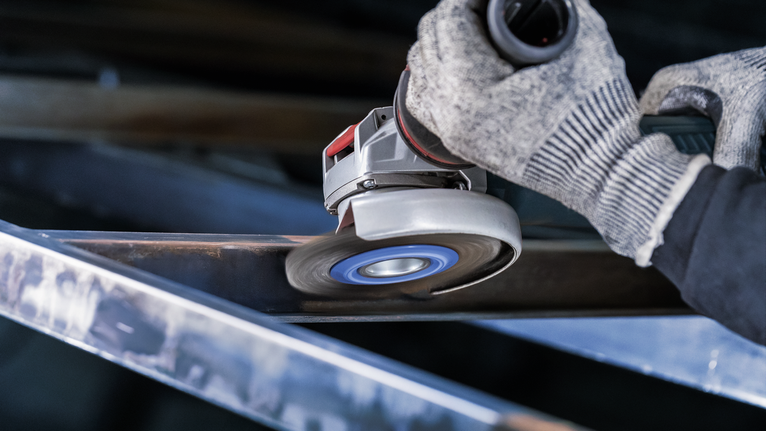 PRO Stainless Steel heavy X-Lock Wire Wheel, 115 x 0.5 mm Person wearing safety equipment grinds a metal beam with an angle grinder.