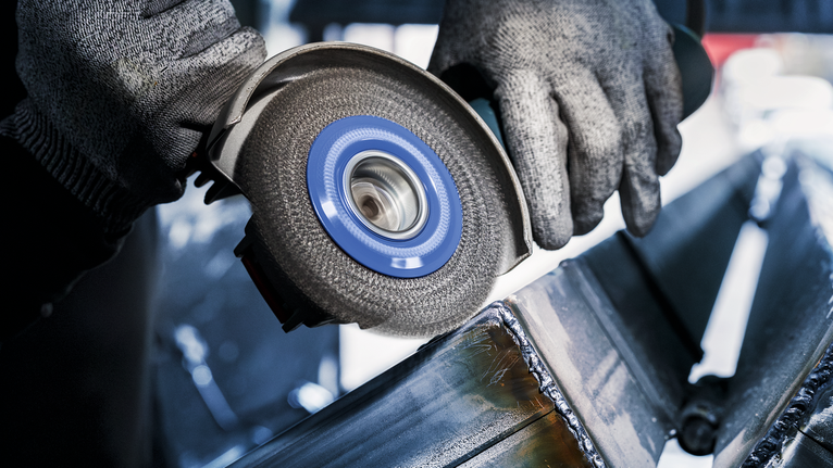 PRO Stainless Steel heavy X-Lock Wire Wheel, 115 x 0.5 mm Person wearing safety equipment grinds welded metal beams with an angle grinder.