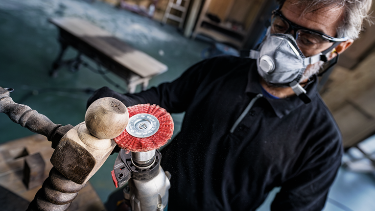 Person wearing safety equipment polishing a wooden spindle with a power tool.
