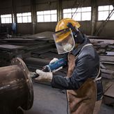 A person wearing safety equipment grinds a metal pipe flange with a straight grinder.