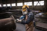A person wearing safety equipment uses a straight grinder to work on a large metal pipe.