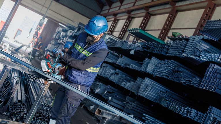 A person wearing safety equipment cuts metal tubing in a warehouse.