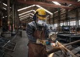 A person wearing safety equipment grinds a metal pipe with an angle grinder in a factory.
