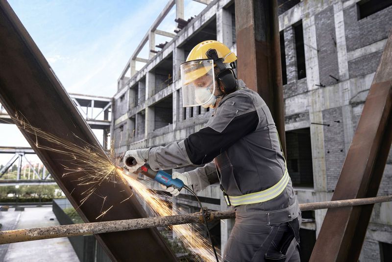 A person wearing safety equipment uses an angle grinder to cut a steel beam.