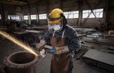 A person wearing safety equipment grinds metal with a power tool in a factory.