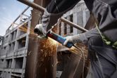 A person wearing safety equipment grinds a steel beam, producing bright sparks.