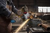 A person wearing safety equipment grinds metal pipe with an angle grinder, producing sparks.