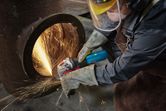 A person wearing safety equipment grinds the edge of a large metal pipe, creating sparks.