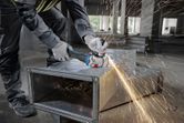 A person wearing safety equipment grinds a metal duct, creating sparks in a construction area.