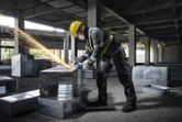 A person wearing safety equipment uses an angle grinder on a metal duct in a construction site.