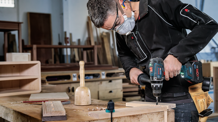 Person wearing safety equipment drills into wood on a workbench.