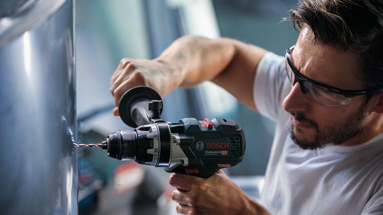 Person wearing safety equipment drills into metal with a power tool.