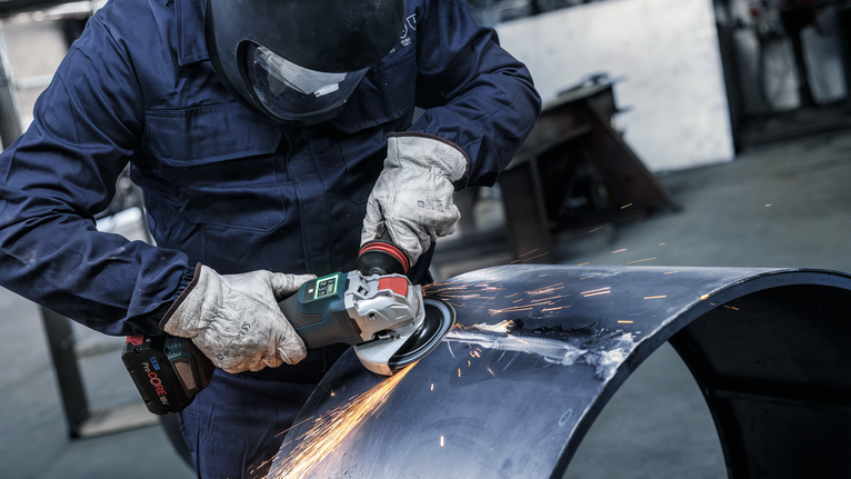 A person wearing safety equipment grinds metal pipe, creating bright sparks.
