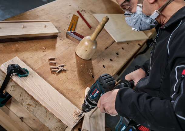 PRO Wood Brad Point Drill Bit Set, 8 pcs Person wearing safety equipment drills a hole in wooden boards on a workbench.