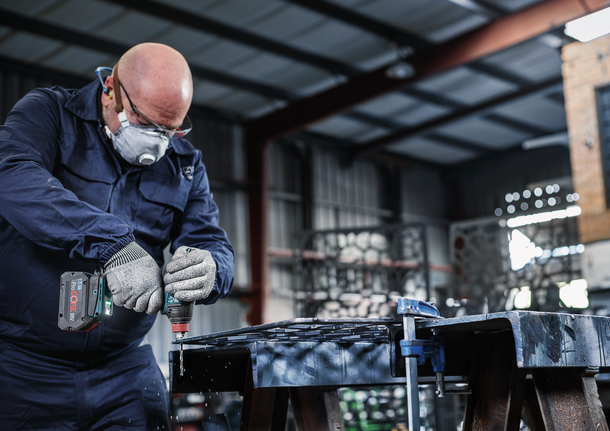Person wearing safety equipment drills a hole through metal in a workshop.