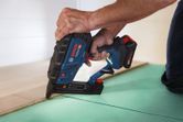 A person uses a cordless wood stapler to fasten wood flooring along a green underlayment.