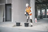 A person wearing safety equipment uses a stirring mechanism to mix material in a bucket.