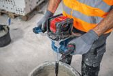 A person wearing safety equipment uses a stirring mechanism to mix material in a bucket.